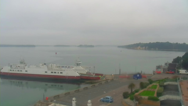 A foggy waterfront scene featuring a large ferry docked at a pier. The ferry has a white body with red accents and displays safety signs. In the foreground, there is a paved area and some greenery with manicured landscaping. Several vehicles, including a blue car and a red car, are parked nearby. In the background, the calm water extends to distant green islands, all under a cloudy sky.