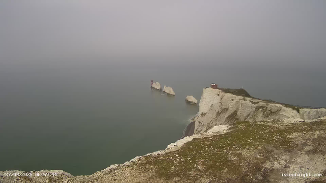 A foggy coastal scene featuring a rocky cliff edge with patches of grass. In the distance, several large, white rock formations rise out of the water. A red structure is visible on one of the rocks, while another small building is perched atop the cliff to the right. The water appears calm and grey, blending into the misty sky, creating a serene but obscured atmosphere.