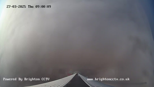 A wide-angle view of a cloudy sky, with thick gray clouds dominating the scene. The foreground features a sloped rooftop, leading towards the center of the image. The timestamp at the top indicates it is Thursday, March 27, 2025, at 9:00 AM. The bottom of the image displays the text "Powered by Brighton CCTV" and the website URL "www.brightoncctv.co.uk". Overall, the scene appears gloomy and overcast.