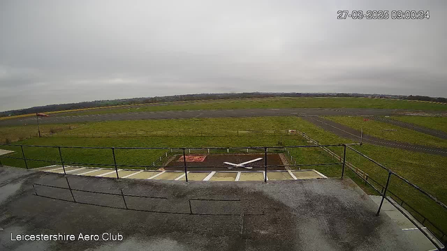 A wide view of an airport runway taken from a webcam. The scene shows a grassy area surrounding the runway, which is mostly clear with a few marked lines. In the foreground, there is a landing pad marked with a white cross and a red area nearby. A wooden fence lines the perimeter, and a flagpole is visible with a flag flying in the wind. The sky is overcast with grey clouds. The image is timestamped at 09:00 on March 27, 2025.