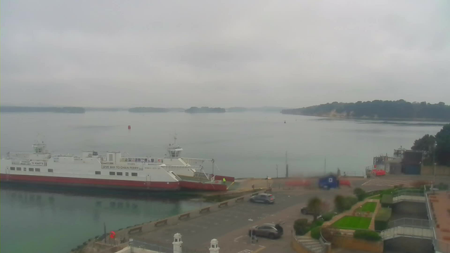 A view of a calm harbor on a cloudy day. In the foreground, a large white ferry with a red stripe is docked at the edge of the water. The boat has a deck area visible. To the right, there is a road with parked cars and a small blue structure. Beyond the ferry, the water is smooth and reflects the sky, with several green islands in the distance. The scene has a tranquil, overcast atmosphere.