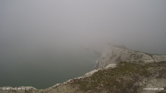 A foggy coastal scene with cliffs stretching into the distance. The view is hazy and obscured, making it difficult to see details. The shoreline appears rocky with patches of grass visible at the edge of the cliff, and a calm sea below. No distinctive features are discernible due to the fog.