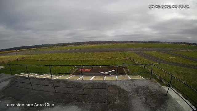 A view from a webcam showing a vast outdoor area. In the foreground, there is a gray rooftop with a railing. Below, a grassy landing area features a white X marking on the ground. The background has an airstrip leading into a wide expanse of grassy fields. The sky is overcast and gray, indicating a cloudy, potentially rainy day. The time displayed is 09:59:53 on March 27, 2025.