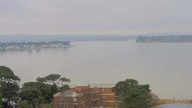A tranquil view of a calm body of water under a cloudy sky. In the foreground, there are several trees and a low brown building with multiple windows. The water reflects the gray sky, and in the distance, there are boats docked by a shoreline lined with trees. The horizon features a gentle slope of green hills, fading into the distance.