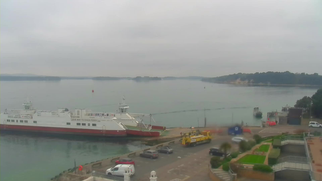 A view of a calm harbor under a cloudy sky. In the foreground, a large white and red ferry boat is docked at the pier. Next to it, there is a yellow construction vehicle near parked cars. Across the water, small boats can be seen, while distant land is visible on the horizon. The scene has a muted color palette due to the overcast weather.