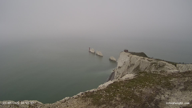 A foggy coastal scene features white chalk cliffs on the right side of the image, with green grass and rocky terrain in the foreground. In the distance, there are jagged rock formations emerging from a calm sea. A small red lighthouse is positioned near the rocks, and a structure is visible at the top of the cliffs. The sky is overcast, blending into the sea. A few people can be seen walking along a pathway at the edge of the cliffs.