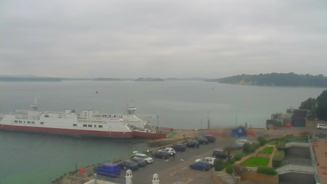 A view of a harbor under a gray sky. In the foreground, a large ferry boat is docked by the water, with a white body and a red bottom. There are several parked cars lined up along the waterfront, with a few people visible near the ferry. The background features calm waters with a few small islands in the distance, while the shoreline is lined with trees and buildings. The overall atmosphere is cloudy and serene.