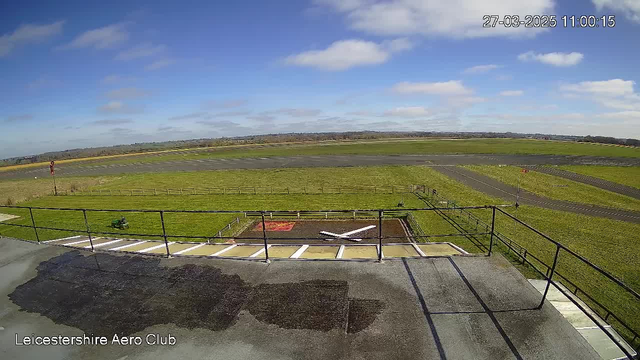 A clear blue sky with a few white clouds is visible above a green landscape. In the foreground, there is a flat rooftop with a ledge and a rectangular helipad marked with a white cross on dark soil. Below the helipad, an open grassy area leads to a runway that extends deep into the background. A red windsock stands on the right side of the image near the edge of the runway, and a wooden fence runs along the grassy field. The scene is bright and sunny, indicating a clear day.