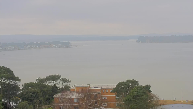 A cloudy sky is visible above a calm body of water, with faint land on the horizon. In the foreground, there are several buildings with brown exteriors and greenery surrounding them. A pathway runs along the water's edge, with a small figure walking in the distance. The scene is tranquil, with a muted color palette of gray and soft earth tones.