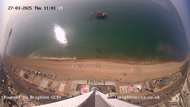 Aerial view of a beach scene featuring a sandy shoreline. The water is calm, with a reflection of sunlight shimmering on the surface. An old pier extends into the sea, and people can be seen walking along the beach. Colorful patterns and structures are visible on the sand. The date and time are displayed in the upper left corner, indicating March 27, 2025, at 11:01 AM. The image is powered by Brighton CCTV.