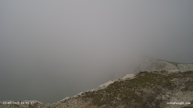 A foggy landscape is depicted, with a rocky foreground featuring patches of grass and small stones. The background is obscured by dense fog, making it impossible to see more than a few meters ahead. The overall scene conveys a sense of emptiness and stillness, as visibility is significantly reduced. The date and time are displayed in the lower left corner.