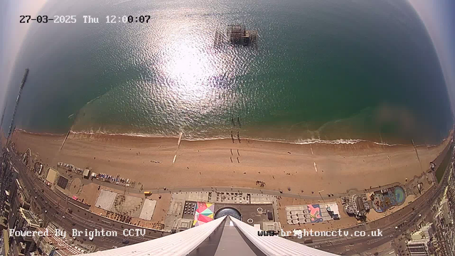 An aerial view captures a wide sandy beach with gentle waves lapping at the shore. The ocean reflects sunlight, creating a shimmering effect on the water's surface. A small, distant pier extends into the sea. Below the beach, a promenade features colorful umbrellas and structures. There are few people walking along the beach and promenade, indicating a quiet day. The scene conveys a peaceful coastal atmosphere. The date and time are displayed at the top of the image.