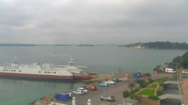 A harbor scene under a cloudy sky. A large white and red ferry is docked at the pier, with a few people in bright clothing visible on board. In the foreground, there is a parking area with a mix of cars, including a blue vehicle and an orange vehicle. The water is calm with a few small islands visible in the distance. On the right side, there are some buildings and greenery, including a landscaped area with paths and planters. The atmosphere appears tranquil and slightly overcast.