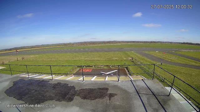 A clear blue sky with some clouds is visible above a grassy landscape. In the foreground, there is a flat surface marked with a white 'X'. A railing surrounds the edge of the platform, overlooking a runway area with asphalt and grassy borders. The distant horizon features a slight rise in land. The scene is bright and appears to be a daytime view from the Leicestershire Aero Club.