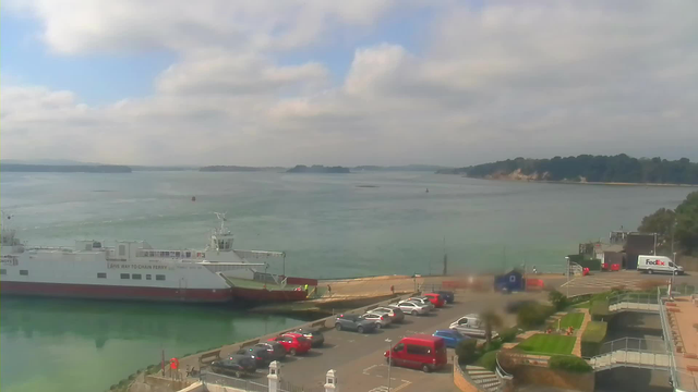 A coastal scene with calm waters and a cloudy sky. In the foreground, a large white ferry is docked at a terminal, partially obscured by a parking lot filled with cars of various colors. To the right, there is a FedEx truck, and small figures can be seen walking near the dock. The background features green hills and trees along the shoreline, with a light breeze causing gentle ripples in the water.