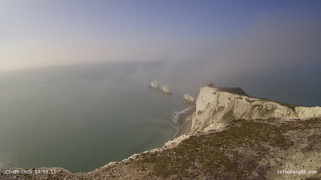 A view of a coastal landscape featuring white chalk cliffs on the right side, with some grassy areas and rocky terrain in the foreground. The ocean extends into the distance, where it meets a hazy sky. In the water, several jagged rock formations are visible, with a lighthouse structure on one of them. The scene depicts a serene atmosphere, with light colors dominating the sky and water. A timestamp is displayed in the lower left corner, indicating the date and time.