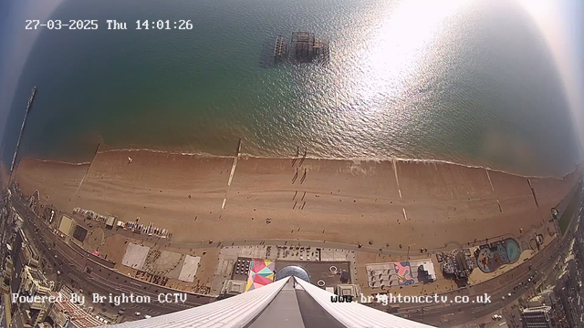 A bird’s-eye view of a sandy beach during daylight, with a calm sea reflecting sunlight. In the background, there is a partially submerged pier structure in the water. The beach is lined with a boardwalk and features scattered figures of people enjoying the day. Colorful beach umbrellas and rides can be seen near the shoreline, along with structures along the promenade. The date and time are displayed in the top left corner.