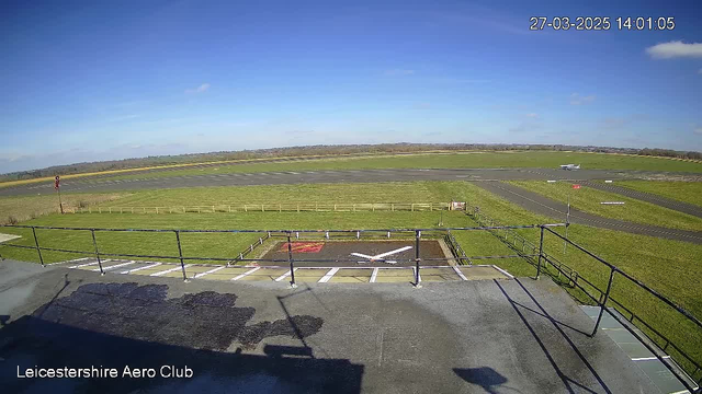 A clear blue sky is visible, with a few scattered clouds. Below, there is a landing strip and grassy area, part of the Leicestershire Aero Club. In the foreground, there is a helipad marked with a large white cross. To the right, an airplane is taxiing on the runway. Fencing surrounds the grass area, and a road runs alongside the runway. The scene conveys a tranquil day at an airfield.
