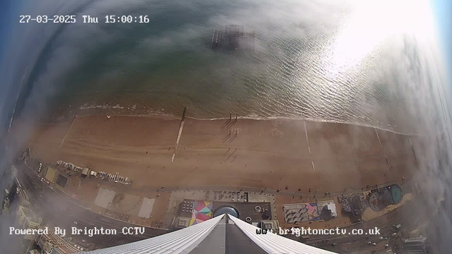 A high-angle view of a sandy beach bordered by a calm sea. The beach is mostly empty, with a few people scattered along the shoreline. In the distance, a pier is partially visible, rising above the water. The image captures a mix of sunlight reflecting off the ocean and some clouds, giving a bright yet slightly hazy appearance. Coastal structures and pathways are visible along the shore, with colorful patterns on the beach near the water's edge. The scene is timestamped with the date and time at the top left corner.