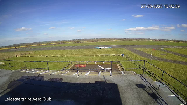 A clear blue sky with a few fluffy white clouds is visible above an airfield. In the foreground, there is a low railing surrounding an area with a red and white helipad marking. To the right, a small plane with a light blue and white color scheme is taxiing on a runway, which is bordered by green grass and a wooden fence. The scene is bright and sunny, showcasing an activity at the Leicestershire Aero Club.