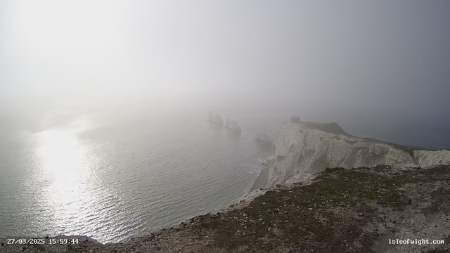 A foggy seascape featuring rocky cliffs on the right side, with several sea stacks emerging from the water in the distance. The ocean appears calm, with a reflective surface. The sky is overcast, blending seamlessly with the fog, creating a muted atmosphere. The foreground shows grass on the cliff edge, leading down to the water.
