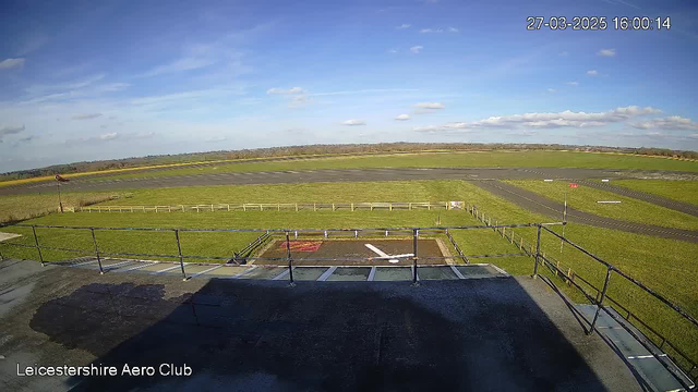 A view from a webcam positioned high above an airfield. In the foreground, there is a ledge with railings. Below the ledge is a helipad marked with a white "H" on a red background. The airfield is visible beyond, featuring a large area of green grass and a gray runway. There is a windsock on the left side, indicating wind direction. In the distance, rolling hills can be seen under a clear blue sky with scattered clouds. The timestamp in the top right corner shows the date and time as March 27, 2025, at 4:00 PM.