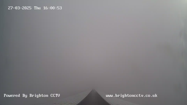 A foggy scene captured by a webcam, with a long, narrow path leading into a dense gray mist. The path is barely visible, disappearing into the fog, which creates a sense of emptiness and obscurity. The top of the image displays a timestamp indicating the date and time. The branding of "Powered By Brighton CCTV" is visible at the bottom.