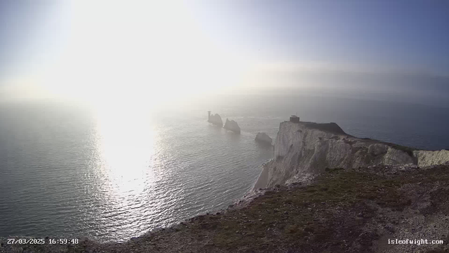 A coastal scene featuring cliffs that drop into the sea. The sun is low in the sky, causing a bright glare on the water's surface. The ocean is calm with gentle waves reflecting the sunlight. Rock formations are visible in the water, and part of a building can be seen atop the cliffs on the right side. The sky is clear with a hint of haze in the distance.