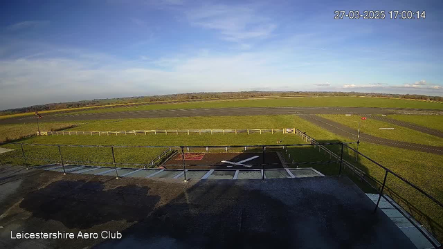 A wide view of an open field at an aerodrome under a clear blue sky. In the foreground, there is a flat rooftop with a railing, and a small area marked with an X shape, likely indicating a landing zone. In the background, the runway stretches across the green terrain, bordered by a white fence. A few clouds are scattered in the sky to the right, and distant hills are visible on the horizon. The time and date are displayed in the top right corner of the image.