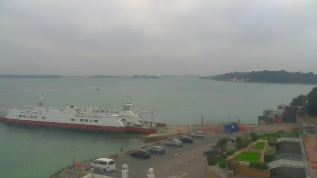 A cloudy waterfront scene featuring a large ferry docked at a harbor. The water is a greenish-blue, with several small islands visible in the distance. In the foreground, there is a parking area with multiple cars, and a well-maintained walkway lined with greenery and landscaping. The sky is overcast, suggesting a gloomy atmosphere.