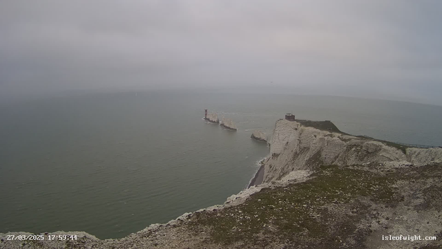 A coastal scene featuring white chalk cliffs and a calm gray sea under a cloudy sky. At the edge of the cliffs, there are patches of green vegetation. In the distance, several rocky outcrops extend into the water, with a lighthouse visible on one of them. The sky is overcast, creating a muted, soft light over the landscape.