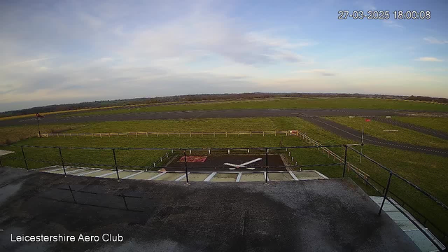 A view from a webcam showing the Leicestershire Aero Club. In the foreground, there is a flat rooftop with a railing. Below, an open grassy area leads to a runway marked with some white lines. To the left, there is a red wind indicator pole. The sky is pale blue with some clouds, and the overall scene is well-lit, suggesting it is late afternoon.