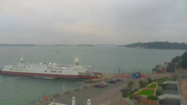 A gray, overcast sky looms over a harbor scene featuring a large white and red ferry docked at the pier. The ferry displays signage indicating it is a chain ferry. In the background, calm waters stretch out toward a distant shoreline with trees. Multiple cars are parked near the ferry terminal, and there is a grassy area with landscaped gardens visible in the foreground. The overall atmosphere is tranquil but somewhat gloomy due to the cloudy weather.
