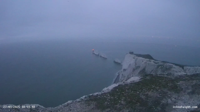 A foggy coastal scene showing a cliffside with rocky outcrops. In the distance, a pier extends into the sea, featuring a small red light. The water is gray and calm under a cloudy sky, creating a misty atmosphere. Limited visibility hints at a tranquil yet eerie setting, with no visible people or boats.