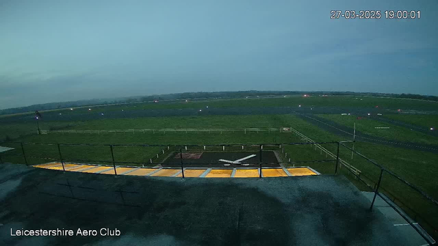 A panoramic view from a webcam positioned at the Leicester Aero Club. The image shows a grassy airstrip under a cloudy sky, with various small lights along the runway. In the foreground, there is a rooftop area with a railing, and orange lighting illuminating the ground. The scene is tranquil and features no visible aircraft, emphasizing the expansive green field of the airstrip.