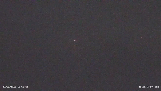 The image is a dark scene with a few faint red and white dots of light scattered across the otherwise black background. The lights appear distant and minimal, indicating a low visibility environment. In the lower left corner, there is a timestamp indicating the date and time of March 27, 2025, at 19:59:42. The bottom right corner features a website link.