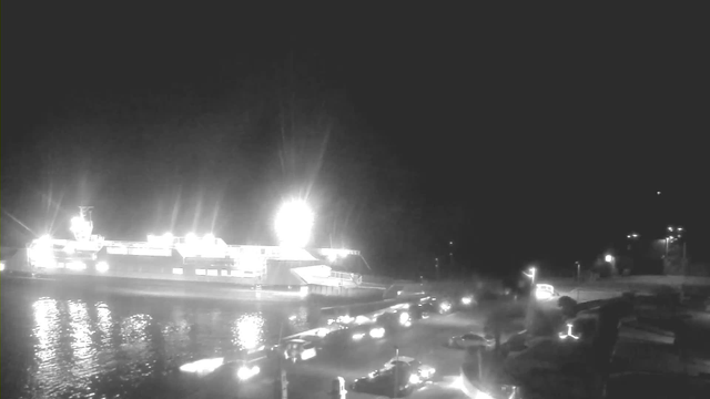 A nighttime scene featuring a large ferry docked at a pier, illuminated by bright lights. The water reflects these lights, creating a shimmering effect. In the foreground, several parked cars line a roadway. Streetlights illuminate the area, providing a faint glow amid the darkness. The background appears to have more cars and possibly additional buildings, though details are less distinct due to the low light.