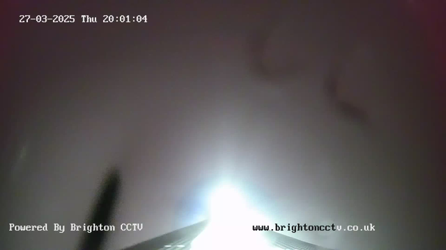 A bright light source is centered in the image, likely representing the sun or a strong artificial light. The background is hazy, appearing foggy or overcast, with minimal visibility beyond the bright source. The date and time are displayed in the upper left corner, indicating March 27, 2025, at 20:01:04. In the bottom left, the text "Powered By Brighton CCTV" is visible, with a website address in the bottom right corner. The overall tone of the image is dim and unclear due to atmospheric conditions.