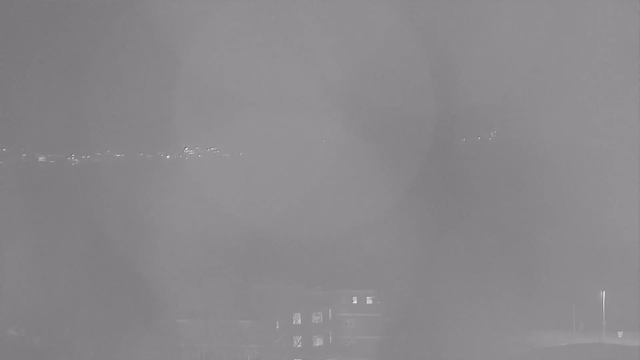 A grayscale image showing a dimly lit scene with some faint lights in the distance, possibly from a cityscape or shoreline. The foreground appears hazy, obscuring details, while a few structures with illuminated windows are partially visible at the bottom of the image. The overall atmosphere is unclear and lacks significant contrast.