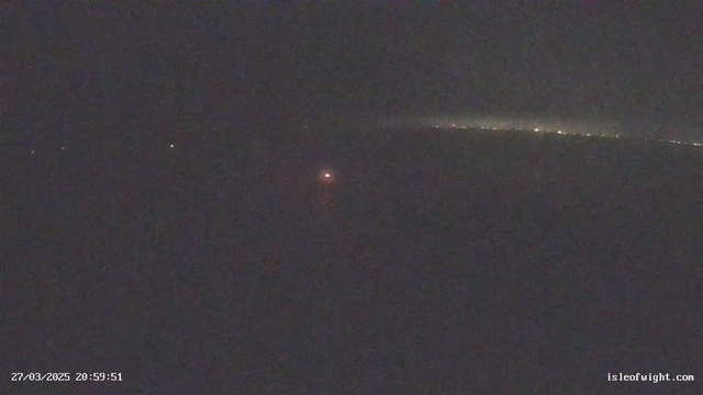 A dark scene with minimal visibility. In the center, there is a faint red light, possibly from a boat or buoy, reflecting off the water. The horizon is barely visible, with a dim line of lights in the distance, suggesting a shore or city. The overall atmosphere is quiet and nighttime, with no discernible details aside from the light and the dark water.
