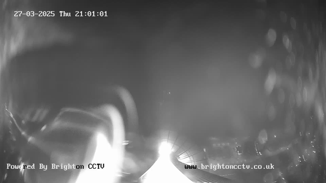 A dark, monochrome webcam image showing a blurred view with bright lights in the center, possibly from headlights or streetlights, illuminating a wide area. The edges of the image are slightly hazy, suggesting possible moisture on the lens. Text at the bottom indicates it is powered by Brighton CCTV, with a timestamp displaying March 27, 2025, at 21:01:01.