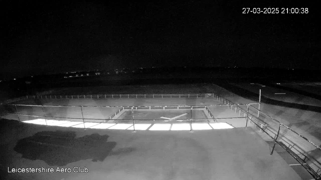 A dark scene depicting an outdoor area of the Leicestershire Aero Club, with a lighted rectangular space in the center that appears to be a helipad. The surrounding area is mostly dim, with a railing visible along the edge of the helipad. In the background, faint lights can be seen at a distance, indicating a possible urban environment. The image is timestamped in the corner, indicating it was captured at 21:00:38 on March 27, 2025.