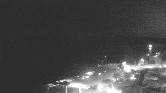 A dimly lit scene shows a waterfront area at night. The horizon is almost entirely dark, with only a few distant lights visible. Closer to the foreground, there are silhouettes of structures and vehicles, with scattered lights illuminating the area. The overall atmosphere is quiet and somewhat mysterious, emphasizing low visibility with limited details.