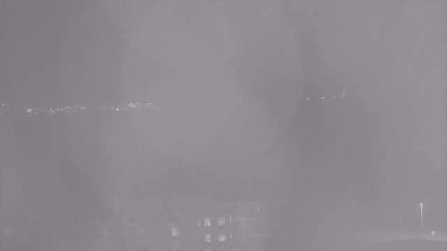 The image is a foggy view captured by a webcam, showing indistinct outlines of buildings and a distant shoreline. A faint line of lights can be seen in the background, likely from structures or vehicles, but overall visibility is very low due to the fog. The foreground is dark, with unclear shapes, and there is a sense of a muted, monochromatic color palette.