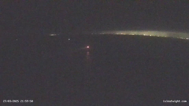 A dark scene depicting a body of water at night. In the foreground, faint reflections can be seen on the surface of the water. To the right, there is a distant coastal shoreline illuminated by scattered lights, creating a soft glow against the dark sky. A single red light appears near the center of the image, possibly from a boat. The date and time are displayed in the bottom left corner.