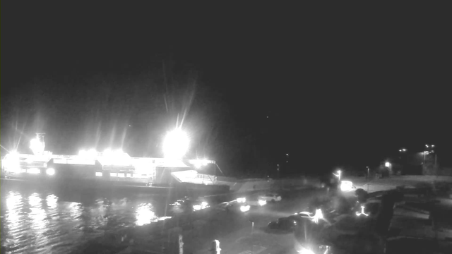 A dimly lit scene of a harbor at night featuring a large ship docked at the water's edge. The ship is brightly illuminated with several lights emanating from its structure, reflecting off the calm water. In the foreground, there are parked vehicles, and additional lights can be seen in the background, likely from nearby buildings or streetlights. The overall atmosphere is dark with prominent lighting from the ship.