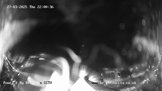 A black and white webcam image showing a blurred view of an illuminated area at night, with streaks of light and dark shapes appearing unclear. At the bottom left, text indicates that the image is powered by Brighton CCTV. The scene seems to depict a cityscape with some lights and possibly a building structure beneath. The date and time are displayed in the upper section of the image.