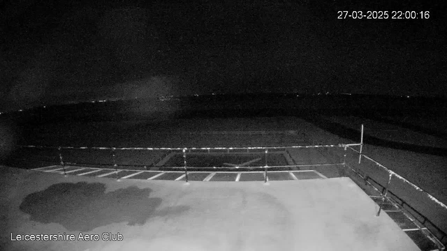 A dimly lit view from a webcam mounted at the Leicestershire Aero Club, showing a flat surface with a large X shape visible on it. The area appears to be mostly dark with some distant lights visible in the background. There is a railing along the edge of the surface, and the sky is mostly black. The image is timestamped for March 27, 2025, at 10:00 PM.