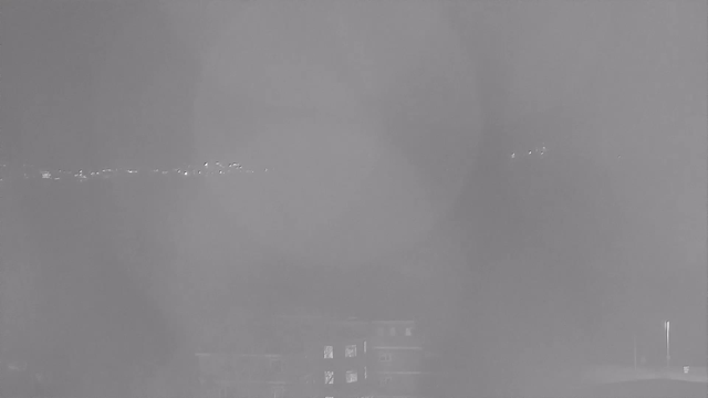 A grainy black and white image taken at night from a webcam, showing a faint horizon lined with small lights in the distance. The foreground is mostly dark, with a blurred shape that resembles a building or structure, and very little detail is visible due to low visibility.