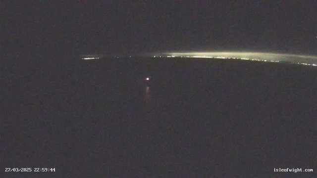 A dark image featuring a small red light near the center, possibly a boat or buoy, reflecting on the water. In the background, along the horizon, there are faint lights indicating the presence of a distant shoreline or city. The overall scene is predominantly dark with minimal light sources.